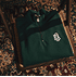 Dunkelgrüner Halfzip Sweater mit FCA-Wappen liegt gefaltet auf einem Stuhl, auf einem gemusterten Te (2)