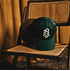 Dunkelgrüne Baseballkappe mit weißem FCA-Wappen liegt auf einem Holzstuhl mit Rattan. (3)